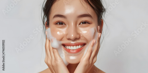 Close-up portrait of smiling asian woman washing face with soap foam, isolated on light grey background