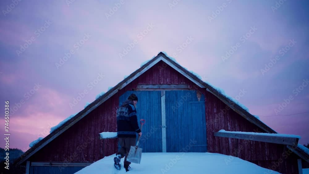 Man With Shovel Walking On Snow-covered Roof Towards Entrance To Attic Of Wooden Cabin At Sunset. wide shot Man With Shovel Walking On Snow-covered Roof Towards Entrance To Attic Of Wooden Cabin At Sunset. wide shot