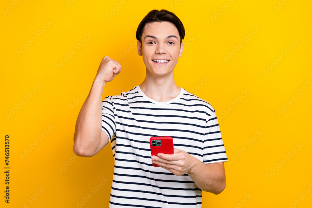 Photo of satisfied guy dressed striped t-shirt hold smartphone clenching fist win gambling isolated on vivid yellow color background Photo of satisfied guy dressed striped t-shirt hold smartphone clenching fist win gambling isolated on vivid yellow color background