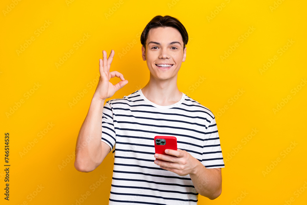 Photo of attractive guy dressed striped t-shirt hold smartphone showing okey approve good quality isolated on yellow color background