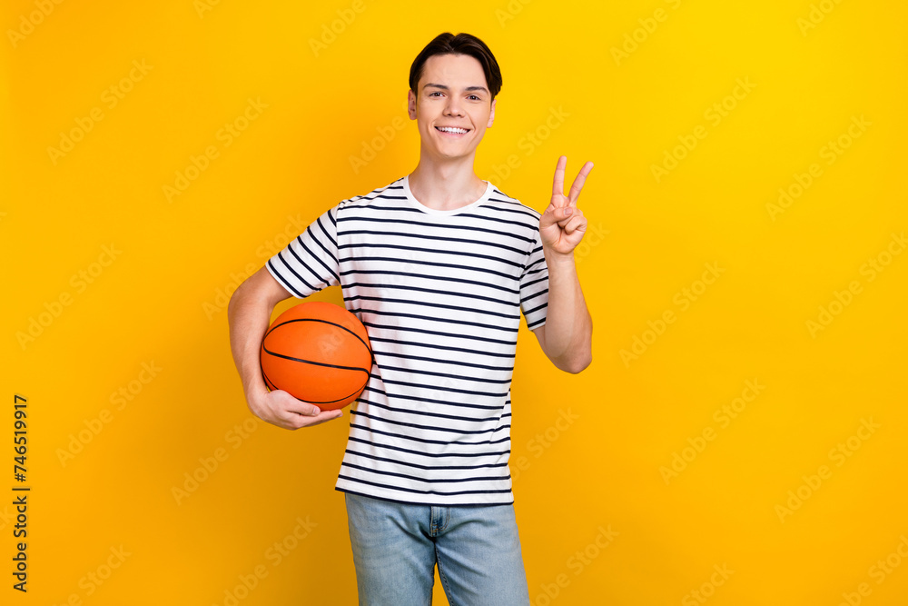 Photo of pleasant student dressed striped t-shirt hold bascketball ball showing v-sign symbol isolated on vivid yellow color background