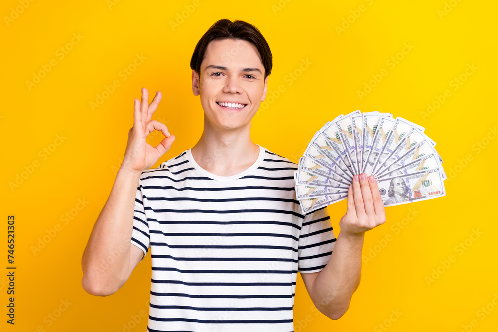 Photo of satisfied guy with brunet hair dressed striped t-shirt holding dollars showing okey nice job solated on yellow color background Photo of satisfied guy with brunet hair dressed striped t-shirt holding dollars showing okey nice job solated on yellow color background
