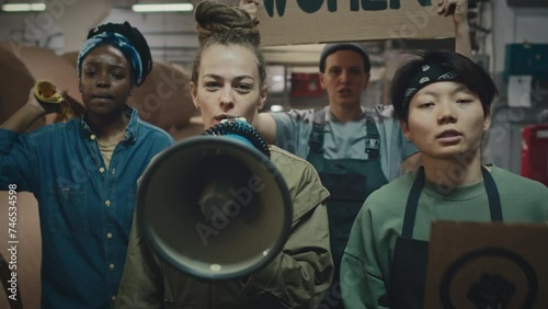 Team of young supportive multiethnic women with mouthpiece and feminist cardboard banners walking along industrial factory and showing their protest determinately photo