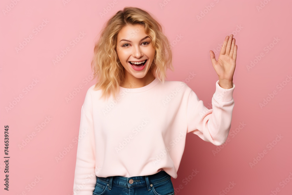 Friendly young woman in a sweater, warmly welcoming with a raised palm and a cheerful smile. Indoor studio portrait on a pink background. Friendly young woman in a sweater, warmly welcoming with a raised palm and a cheerful smile. Indoor studio portrait on a pink background.