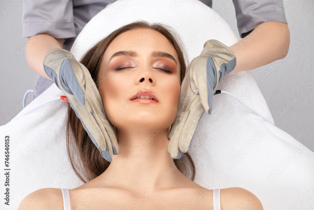 A cosmetologist doctor makes a microcurrent massage procedure on the face and neck of a young beautiful girl in a beauty salon. A cosmetologist doctor makes a microcurrent massage procedure on the face and neck of a young beautiful girl in a beauty salon.