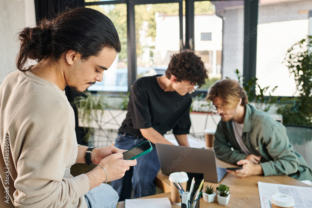 Young professional using smartphone near blurred colleagues brainstorming over a laptop in office