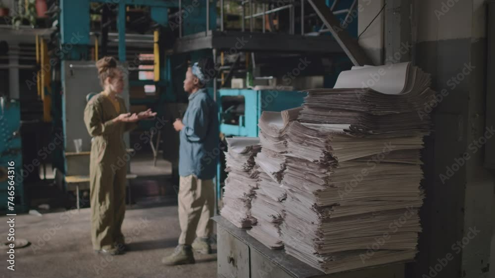 Full shot of two multiracial female employees in workwear having conversation during work shift at printing factory with stacks of paper on old drawer in focused foreground