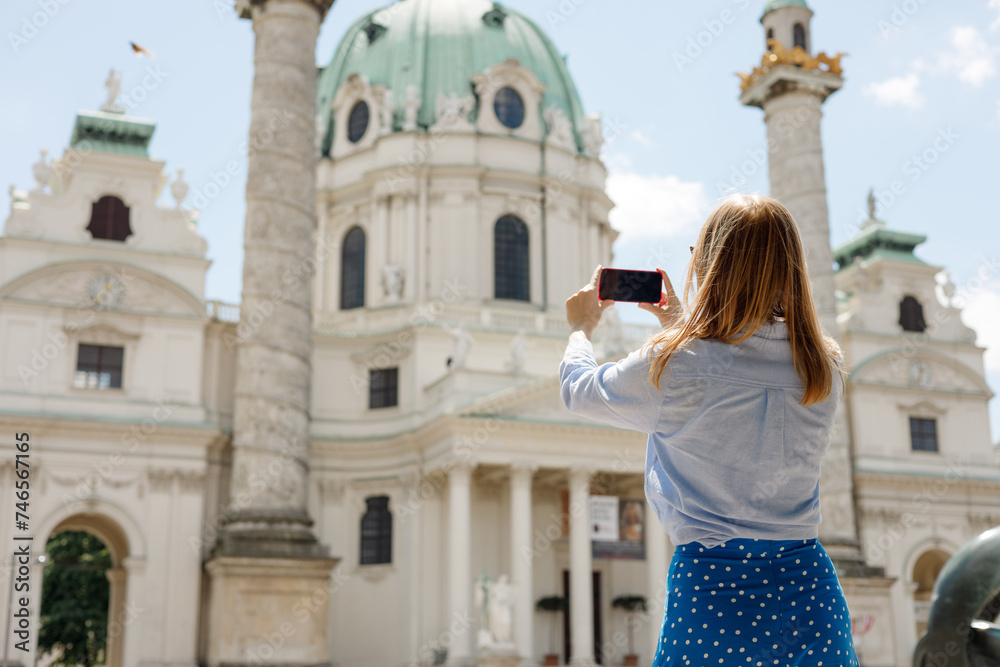 Young female tourist with smart phone. Stylish woman photographing beautiful St. Charles's Church, Vienna. 30s women walking enjoying ancient architecture