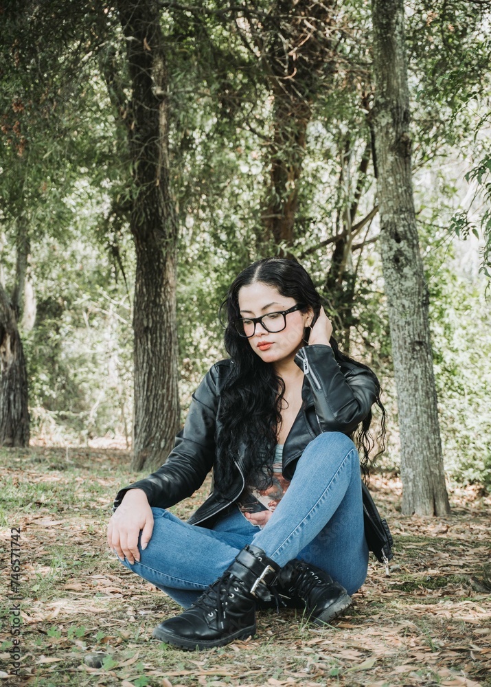 Beautiful latin american brunette woman with black hair, a black jacket and blue pants sitting on the grass Beautiful latin american brunette woman with black hair, a black jacket and blue pants sitting on the grass