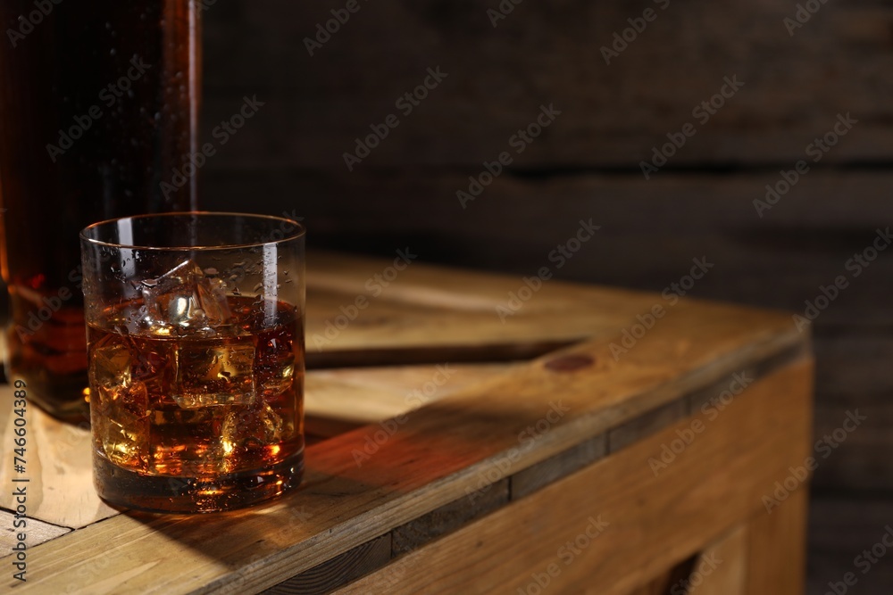 Whiskey with ice cubes in glass and bottle on wooden crate, closeup. Space for text Whiskey with ice cubes in glass and bottle on wooden crate, closeup. Space for text
