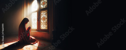 A Muslim woman, covered in a red veil, kneels in prayer as the sun's rays filter through a nearby window. photo