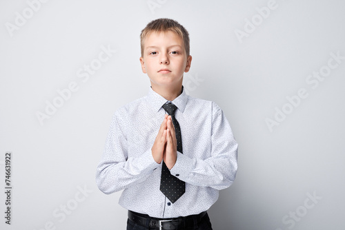 Young boy in shirt and tie with hands together, looking serious on a gray background.