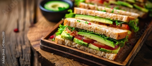 A close-up view of a rustic wooden tray holding delicious vegan club sandwiches topped with fresh avocado slices. The sandwiches are neatly arranged, showcasing the combination of vegan club