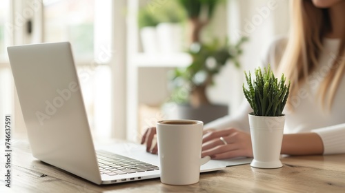 Cropped view of woman sitting at wooden table and working on laptop. A cup of coffee with a potted plant in the background. The environment is bright and clean, a relaxed work environment