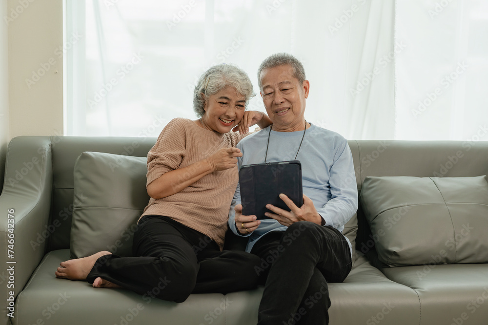 Asian senior couple looking at camera and smiling, senior smiling and looking at tablet Retired people use technology at home Woman hugging man with love Together forever on the sofa at home Asian senior couple looking at camera and smiling, senior smiling and looking at tablet Retired people use technology at home Woman hugging man with love Together forever on the sofa at home