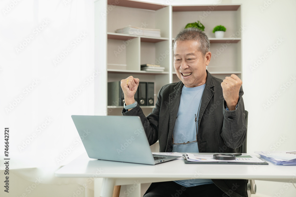 Senior Asian businessman in a suit is excited Happy mature Asian businessman clenching his fist showing victory gesture while sitting on desk in front of laptop at office Senior Asian businessman in a suit is excited Happy mature Asian businessman clenching his fist showing victory gesture while sitting on desk in front of laptop at office