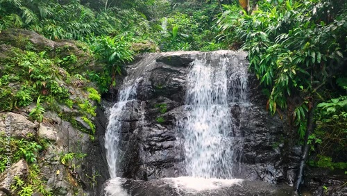 Rising view of a Puerto Rican, tropical waterfall and pool, with lush vegetation and distant background waterfall.