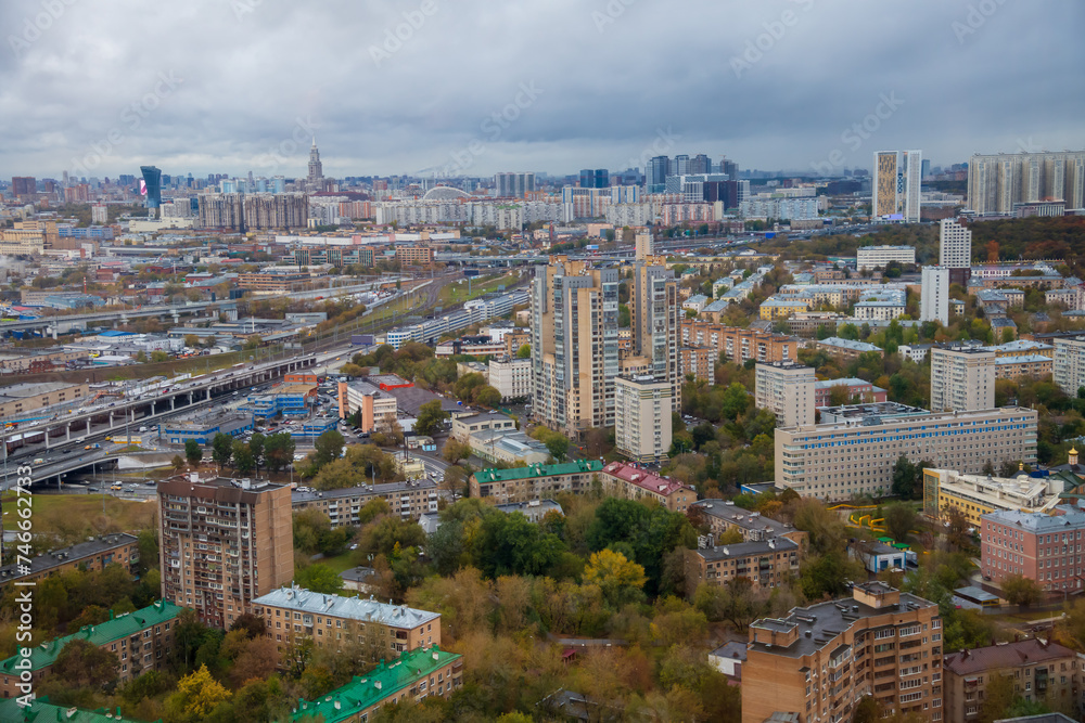High angle cityscape of Moscow city in Russia in a cloudy autumn day. Various residential and industrial buildings next to roads and green and yellow trees. Overcast sky. Soft focus. City life theme.