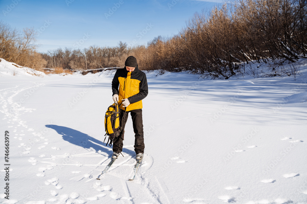Man on skis standing on frozen river with backpack in hands, hiking in winter forest, solo trip.