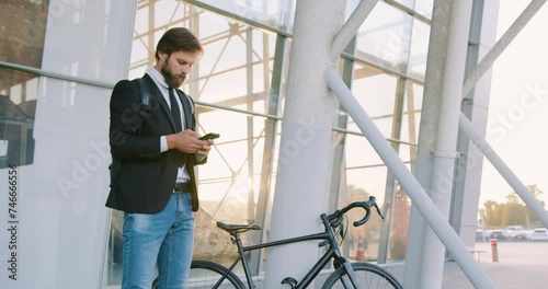 Handsome confident purposeful young business man standing near glass wall with his bike and using smartphone to write message. photo