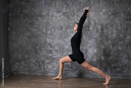 Woman practicing yoga doing Virabhadrasana exercise, warrior pose number one, training in black sportswear in a room near an abstract patterned wall
