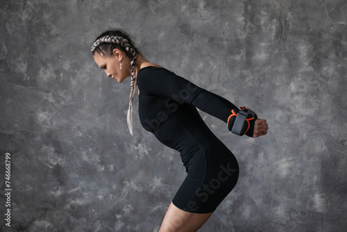 A sporty brunette woman performs exercises with weights on her arms to form a beautiful shape of her arms  stands sideways to the camera near an abstract wall