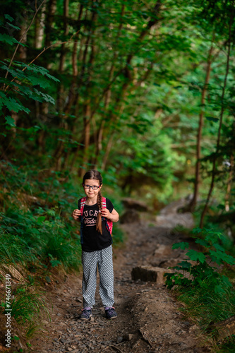 A cheerful young girl with braided hair and glasses embarks on an adventure, hiking a rocky trail in a verdant forest, sporting a casual outfit and a red backpack