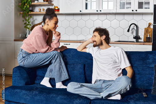 cheerful multiracial couple in homewear sitting on sofa and looking at each other lovingly