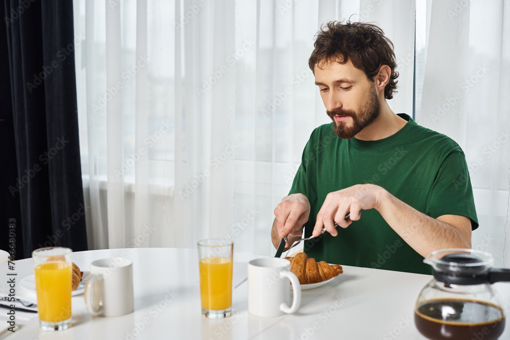 handsome man in casual green t shirt cutting delicious croissant with knife during breakfast at home handsome man in casual green t shirt cutting delicious croissant with knife during breakfast at home