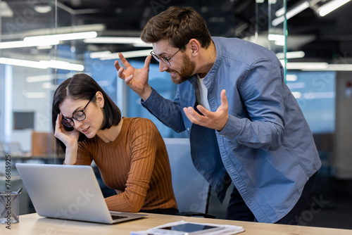 Foto Aggressive male boss in denim shirt spreading hands and yelling at annoyed female employee by working desk