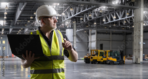 Man in industrial hangar. Warehouse worker with laptop. Contractor inside empty warehouse. Loading equipment behind worker. Man inspects empty hangar. Location of future logistics center