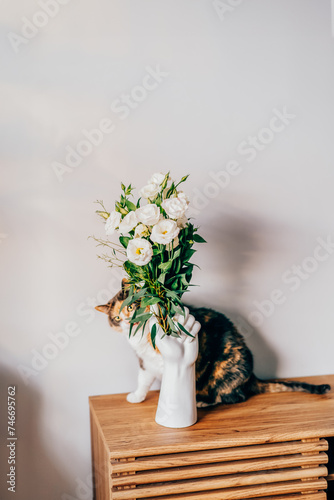 Eustoma flowers bouquet in stylish white ceramic vase made in shape of human hand and multicolored cat pet hiding behind it on wooden console. Trendy interior design decor details. Selective focus.