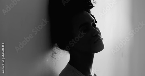 One depressed young black woman leaning on wall feeling lonely in quiet despair struggling with mental illness. Silhouette of African American 20s person suffering alone in black and white photo