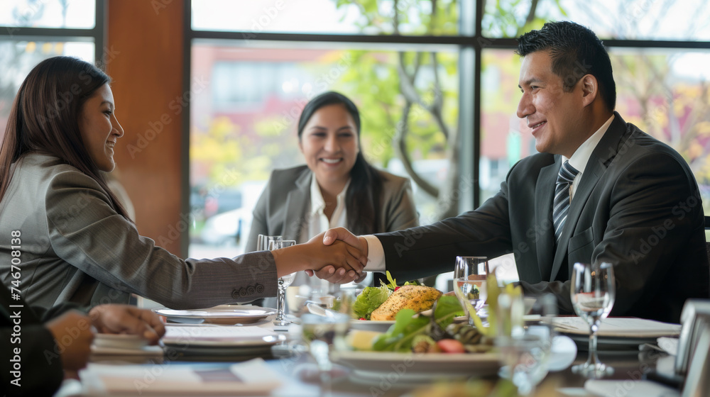 Two professionals are shaking hands across a table in a bright office setting, smiling in agreement or greeting.