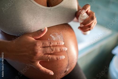 Young woman applying stretch mark cream to pregnant belly at home