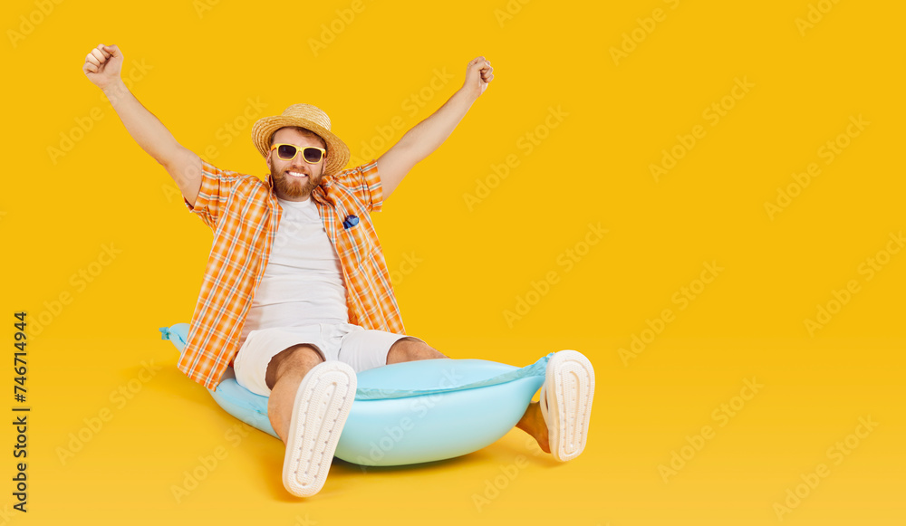 Happy smiling man in sunglasses and hat sitting on inflatable mattress with hands up isolated on a studio yellow background. Bearded guy going to swim on a beach. Vacation and holiday trip concept. - obrazy, fototapety, plakaty 