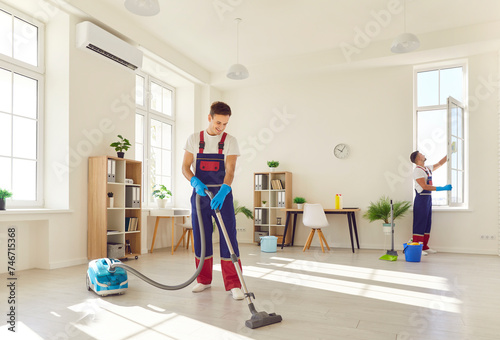 Team of cleaning service workers tidying up office or apartment. Two male janitors working in modern interior. Young man cleaner vacuums floor while coworker washes windows