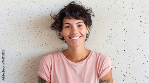 A person with a joyous smile and short hair, featuring a tattoo on their arm and piercings, is wearing a soft-colored shirt against a textured white background.