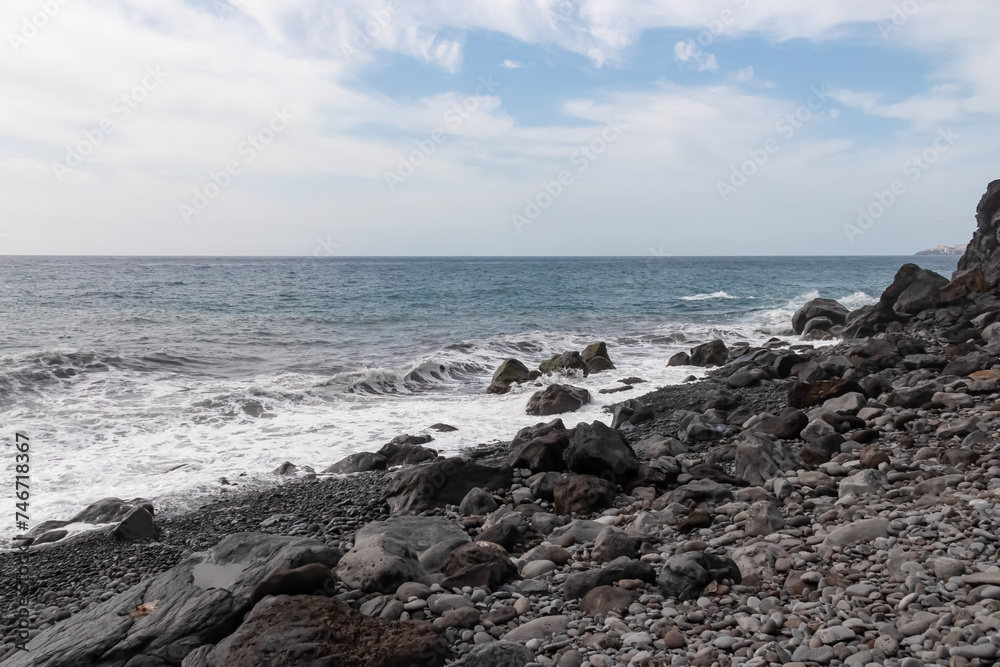 Panoramic view of idyllic volcanic black stone beach of Praia Garajai, Canico, Madeira island, Portugal, Europe. Sea waves hitting shoreline of majestic Atlantic Ocean. Dramatic sky. Coastal landscape
