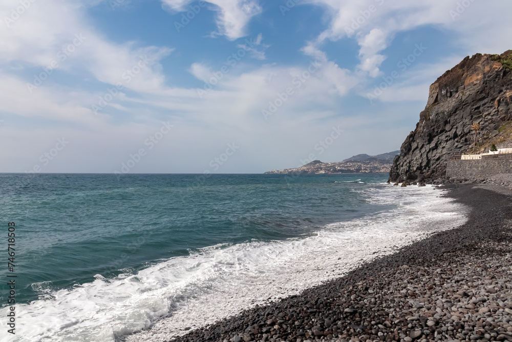 Panoramic view of idyllic volcanic black stone beach of Praia Garajai, Canico, Madeira island, Portugal, Europe. Sea waves hitting shoreline of majestic Atlantic Ocean. Dramatic sky. Coastal landscape