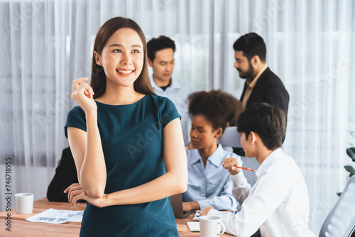 Young Asian businesswoman poses confidently with diverse coworkers in busy meeting room background. Multicultural team works together for business success. Office lady portrait. Concord © Summit Art Creations