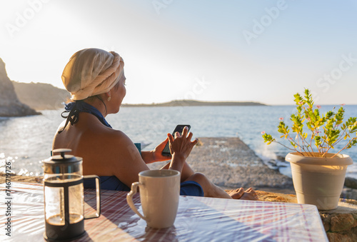 Beautiful woman with Greek outfit on her head and blue sarong checking her mobile phone while having breakfast in a typical Greek tavern by the Aegean Sea in summer