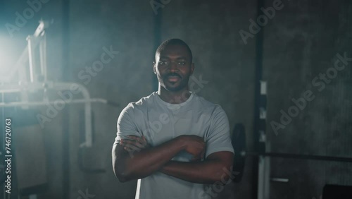 African american sportsman in gym. Man in white t-shirt smiling standing in close pose with hands crossed on chest ready for sport training. Healthy lifestyle, athlete habits, sportive body concept. photo