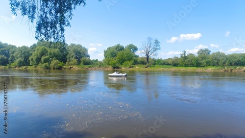 A motorized plastic boat is floating on the river and fishermens are trolling for predatory fish. Willow branches lean over the water. Trees and shrubs grow along the banks of the river. Sunny weather photo