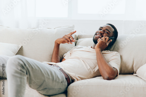 Happy African American man sitting on a black sofa, holding a mobile phone and smiling while making a video call The modern apartment and relaxed atmosphere complement the casual lifestyle and use of