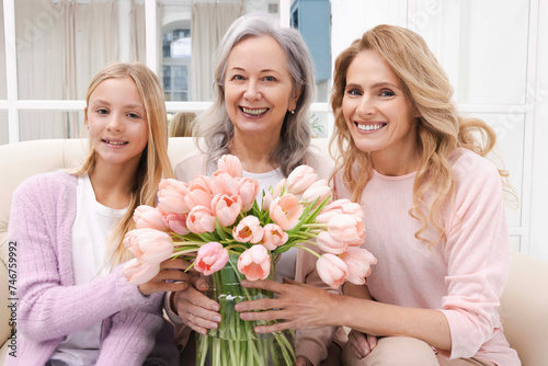 Mother and daughter giving grandmother bouquet of spring flowers, celebrate her birthday and taking photo for memory, sitting on couch