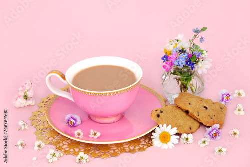 Cup of tea with homemade chocolate chip cookies, Spring flowers and wildflowers in a vase and scattered. Luxury teacup on gold doily on pink background.