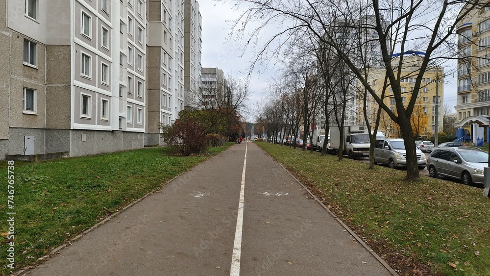 Paved sidewalks and a bike path are laid between the carriageway of the street with parked cars and residential buildings. There are trees on the grassy lawn. Cloudy autumn weather