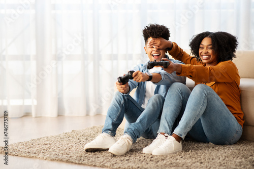 Joyful African American couple playing video games on the floor, with the woman playfully covering the man's eyes #746775709