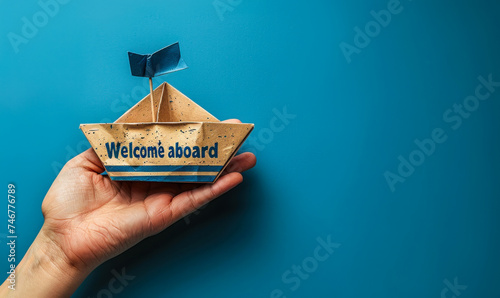 Hand presenting a paper boat with the message Welcome aboard against a blue background, symbolizing new beginnings and joining a team or company photo